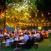 Outdoor evening event with string lights and people seated at tables under trees.